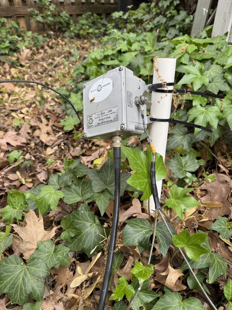 Balun Designs 4:1 Unun 2kW Model 4132sw zip-tied to a PVC pipe hammered into the ground. Here you can see the feed line connecting to the bottom and the radials (3) connected to the side.
